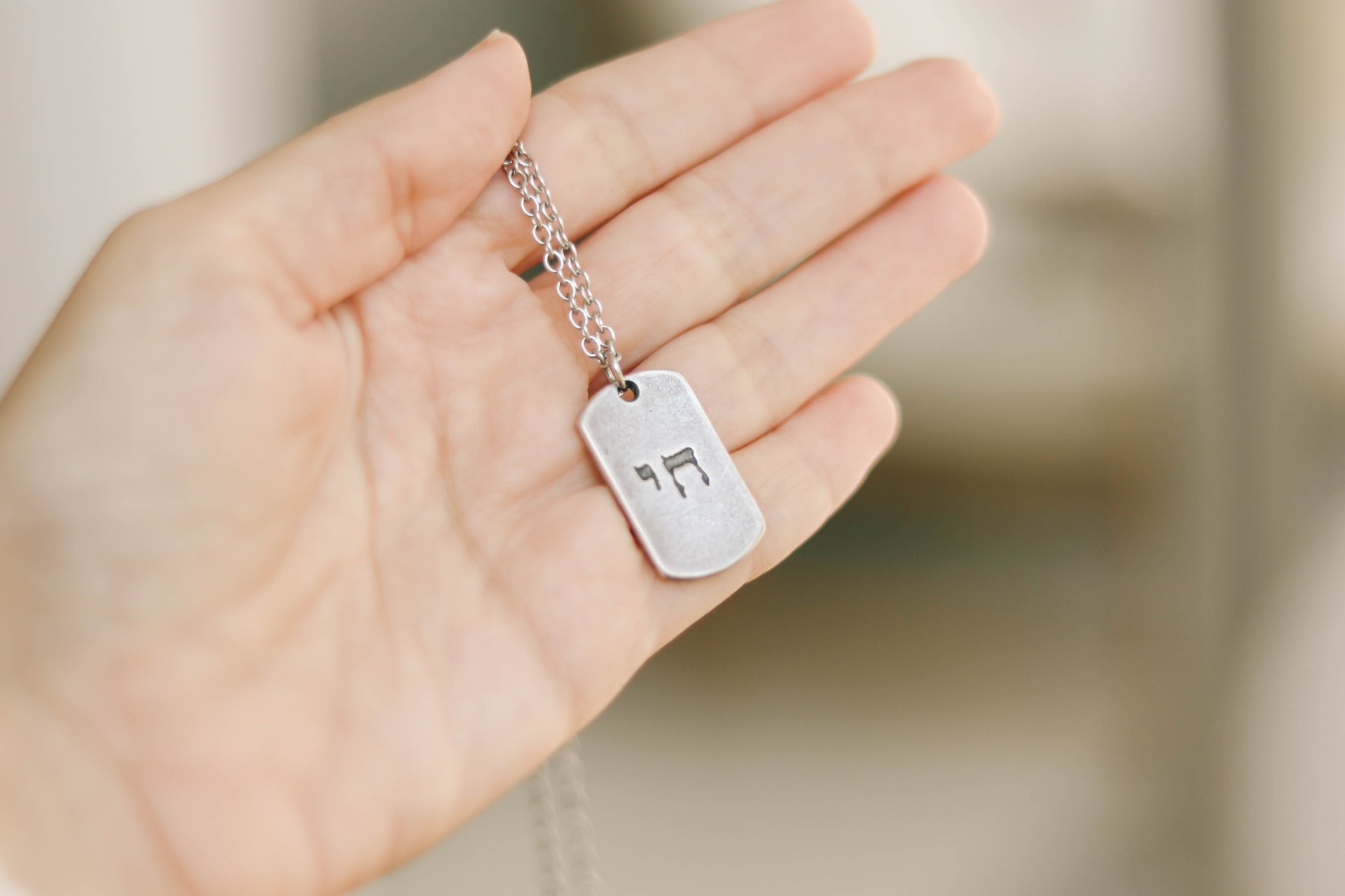Hand holding a silver necklace with a tag featuring Hebrew characters against a blurred background
