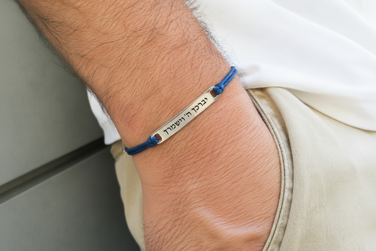 A close-up of a person's wrist wearing a blue cord bracelet with a silver metal plate engraved with Hebrew text.