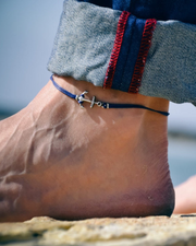 Anchor anklet for men on a foot with jeans rolled up, near water.