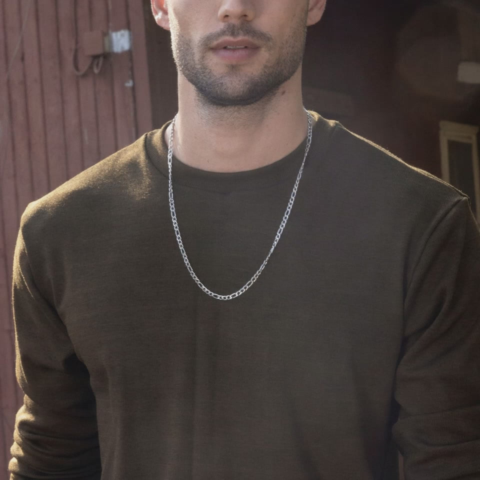 A man wearing a green sweater and brown pants, standing in front of a rustic backdrop.