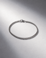 A silver-toned metal chain bracelet with a lobster clasp, displayed on a textured gray surface with dramatic lighting.
