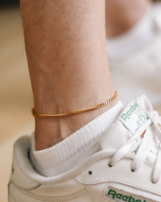 Gold anklet on a person's ankle with a white sneaker and green laces.