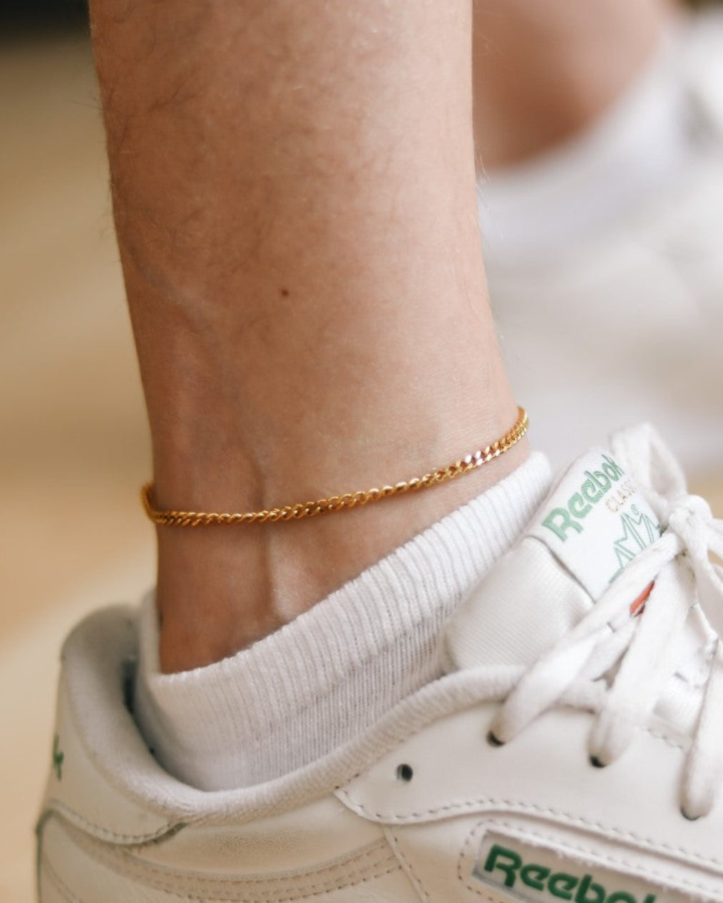 Gold anklet on a person's ankle with a white sneaker and green laces.