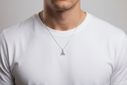 Man wearing a white t-shirt with a silver necklace featuring a triangular trinity pendant on a gray background